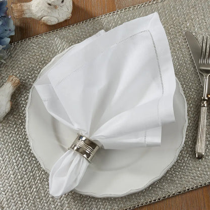 White folded hemstitch napkin with a silver napkin ring on a white plate, placed on a textured placemat with cutlery.