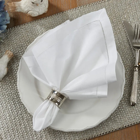 White folded hemstitch napkin with a silver napkin ring on a white plate, placed on a textured placemat with cutlery.
