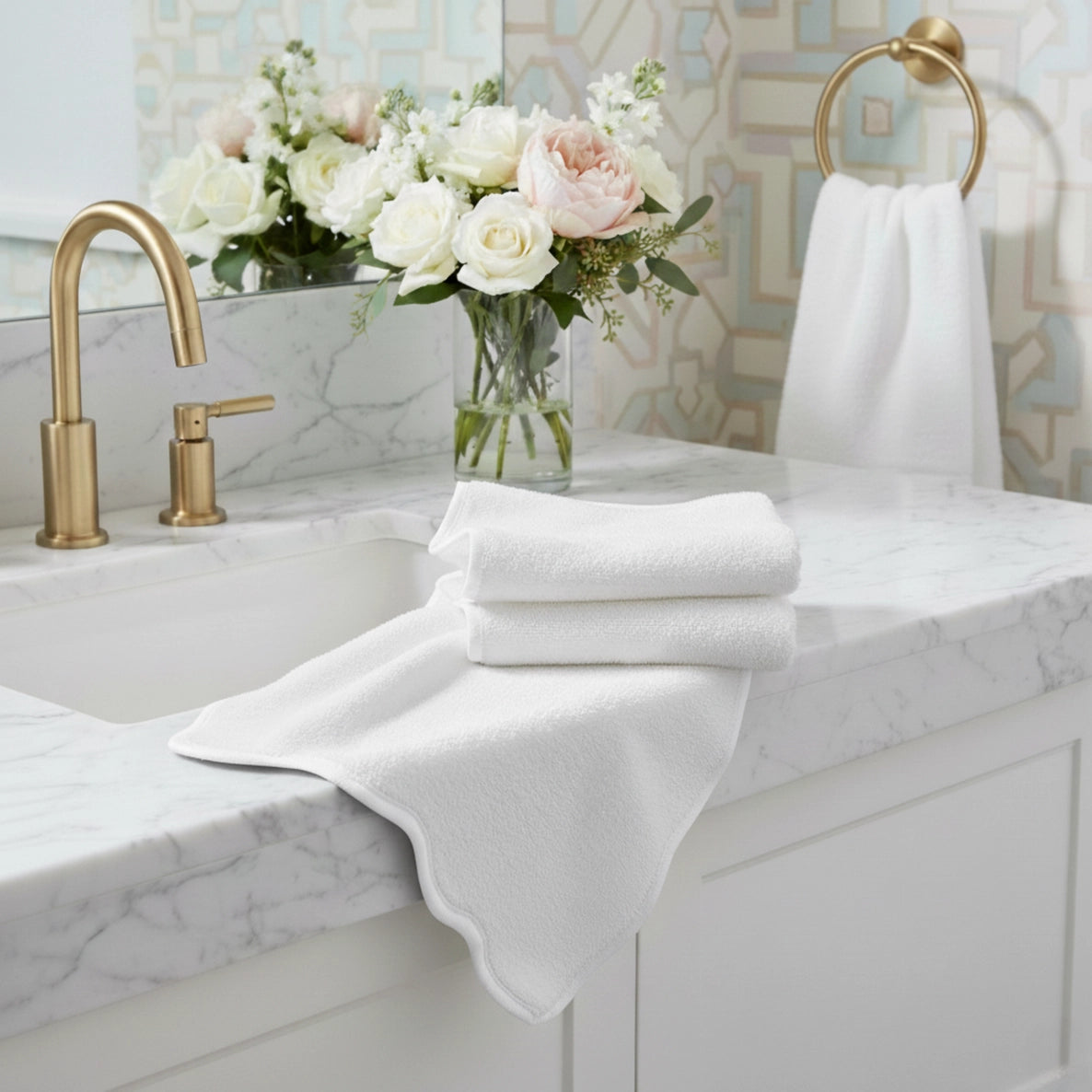 White scalloped edge towels on a marble countertop with a vase of flowers and gold fixtures.